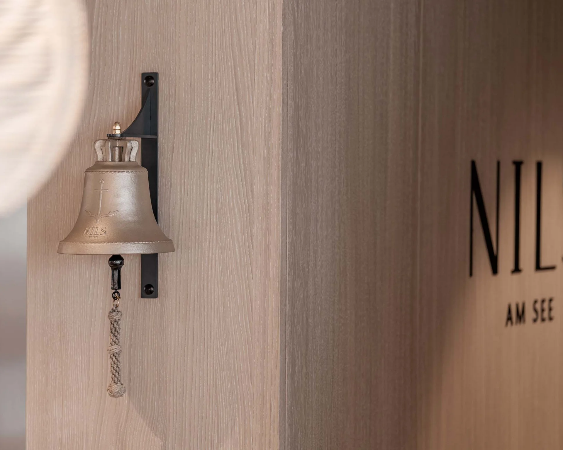NILS am See: Impressionen eines Orts für das süße Leben Metallglocke mit Ankermotiv an Holzwand neben Schriftzug NIL am See