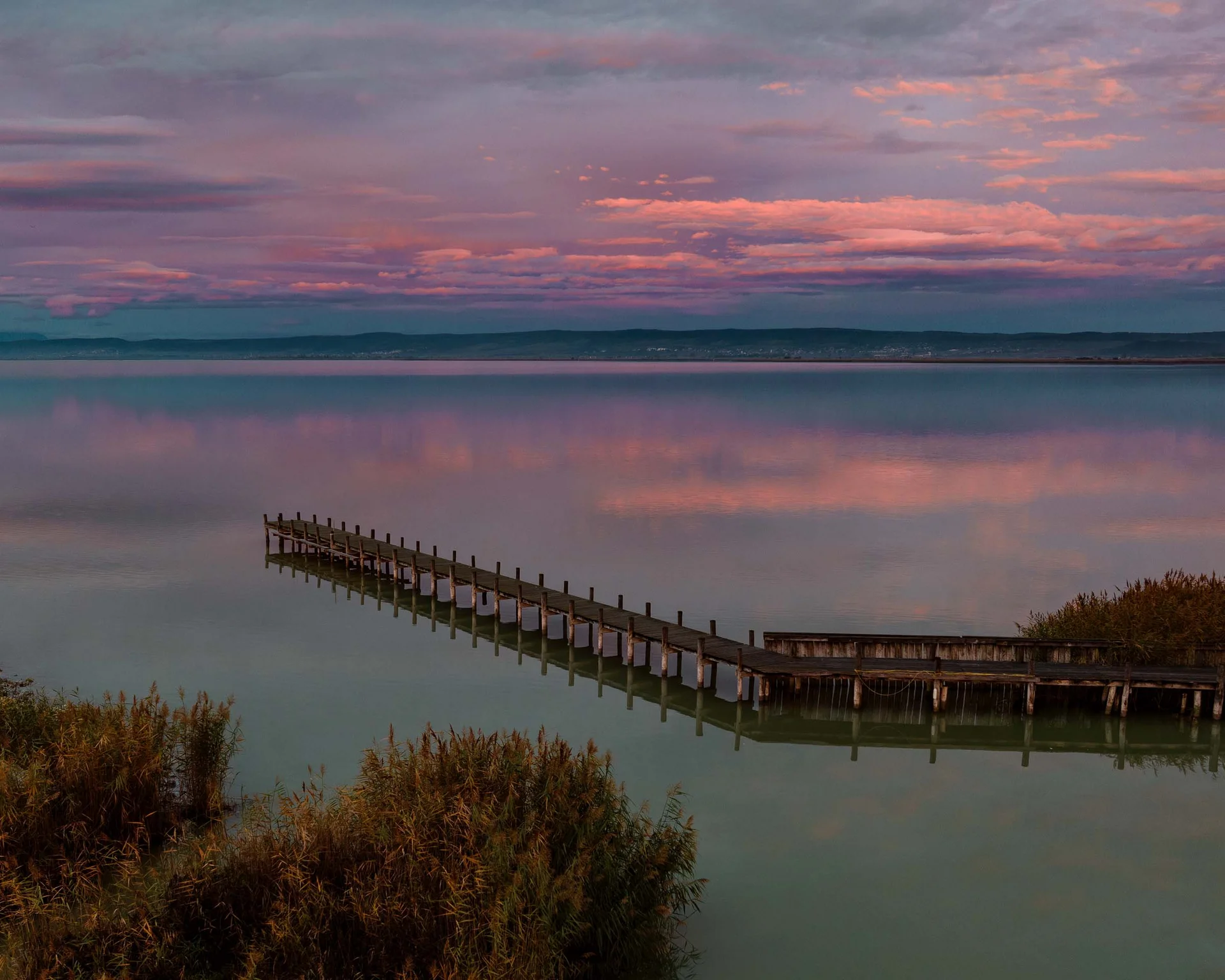 NILS am See: Impressionen eines Orts für das süße Leben Steg auf ruhigem See bei Sonnenuntergang mit rosa bewölktem Himmel