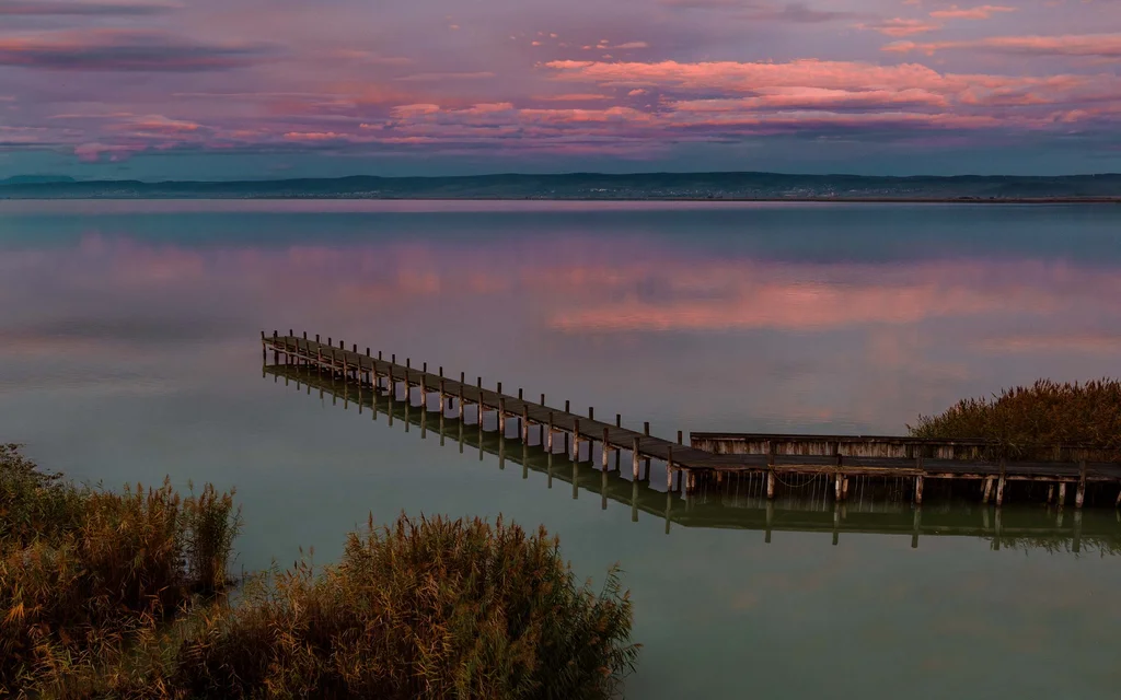 NILS am See: das Hotel am Neusiedler See mit Pool und Seebad Steg auf ruhigem See bei Sonnenuntergang mit rosa bewölktem Himmel