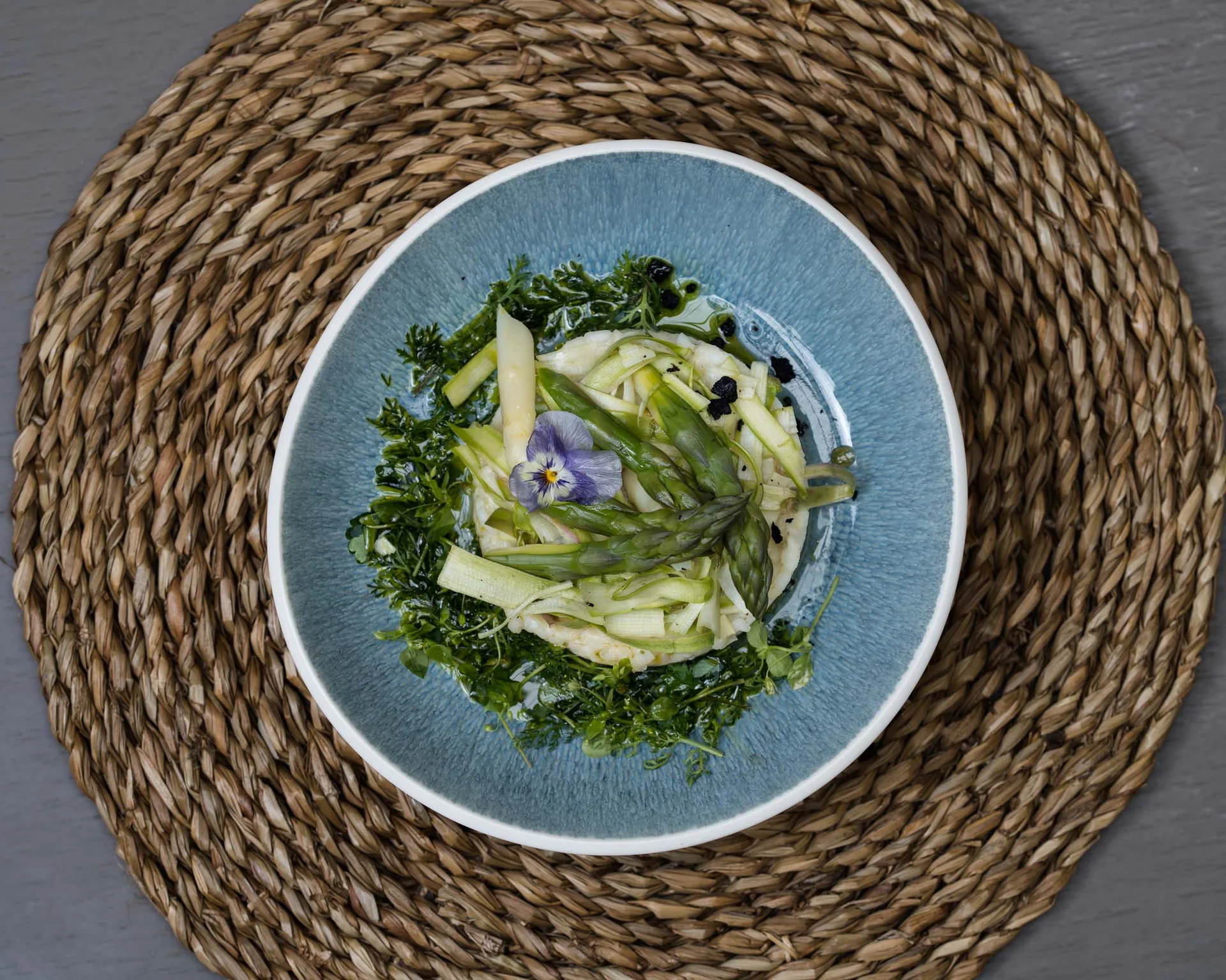 NILS am See: Impressionen eines Orts für das süße Leben Blauer Teller mit grünem Spargel und essbarer Blüte auf geflochtenem Untersetzer