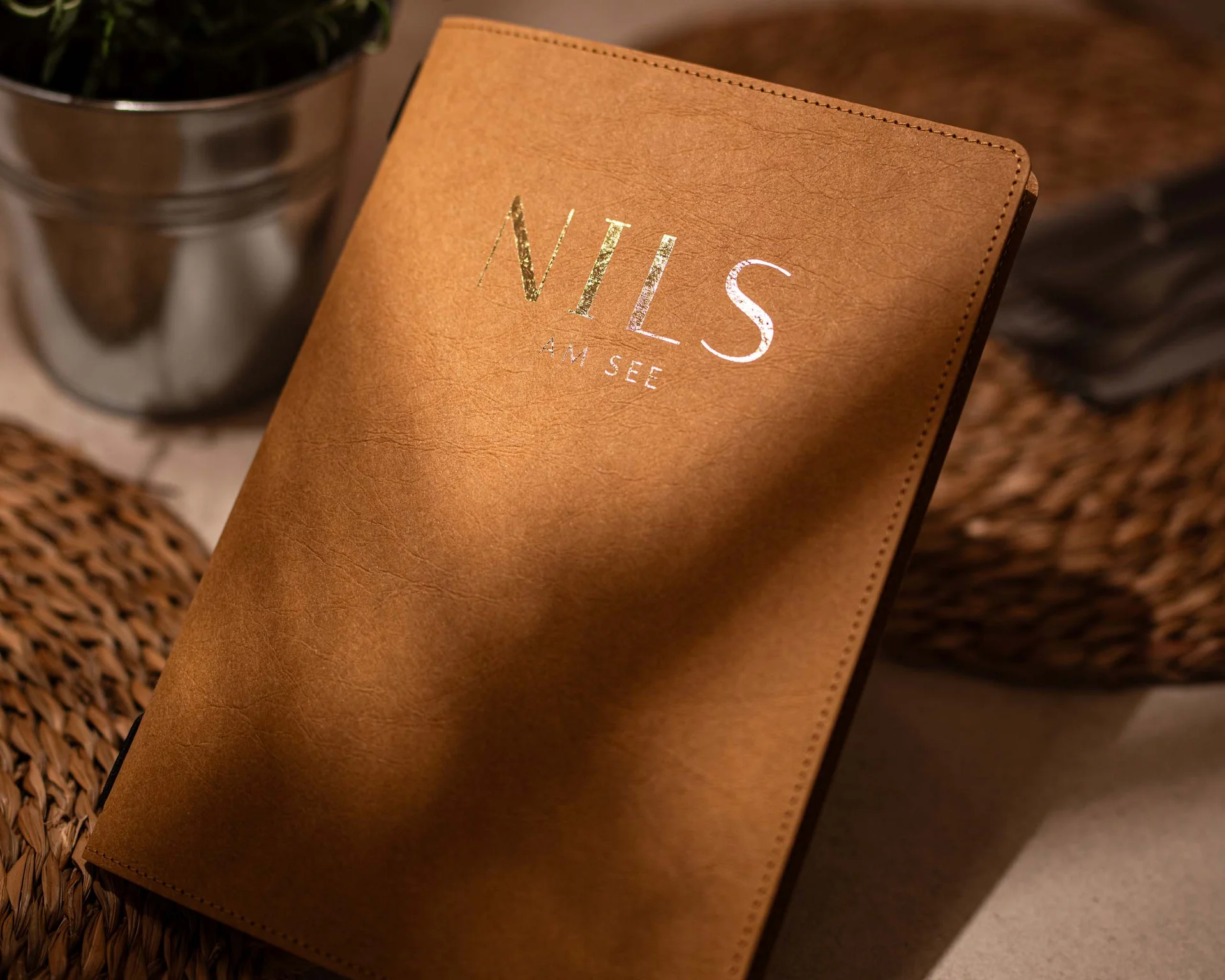 NILS am See: Impressionen eines Orts für das süße Leben Braunes Notizbuch mit der Aufschrift NILS auf einem Tisch neben einem Pflanzenübertopf