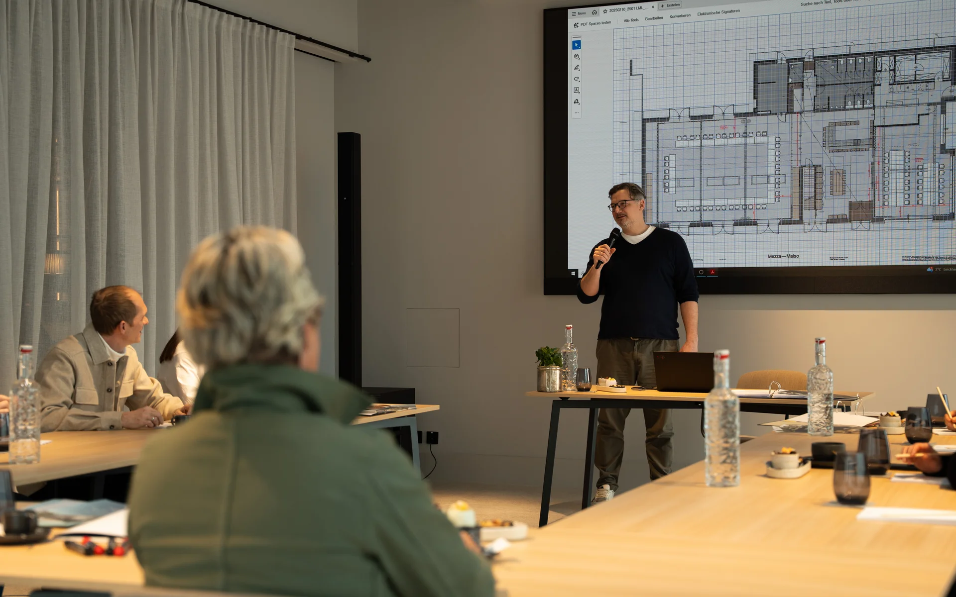 NILS am See: unser Event- und Seminarhotel im Burgenland Person hält Präsentation mit Bauplan vor einem Konferenztisch mit Zuschauern