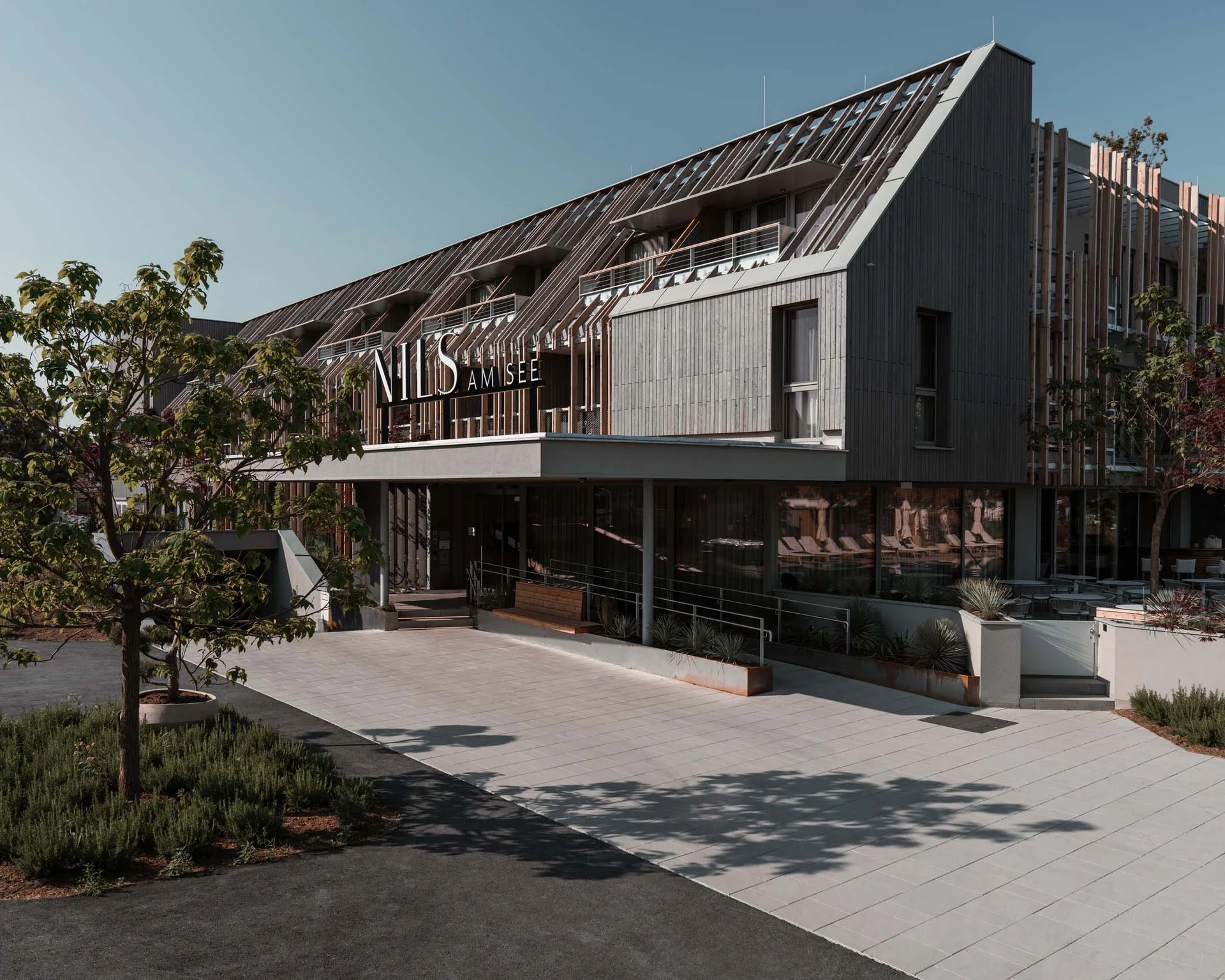 NILS am See: Impressionen eines Orts für das süße Leben Modernes Hotelgebäude mit Holzfassade und Bäumen am Eingang