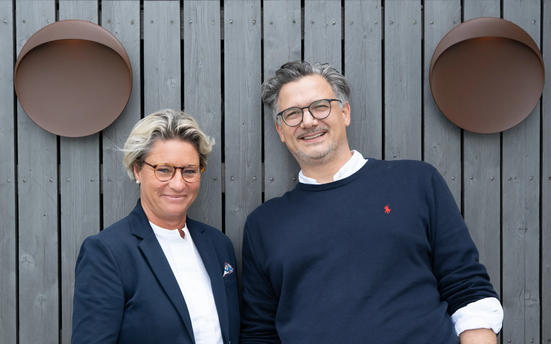 NILS am See: Philosophie und Gastgeber Zwei lächelnde Personen vor einer grauen Holzwand mit runden Wanddekorationen