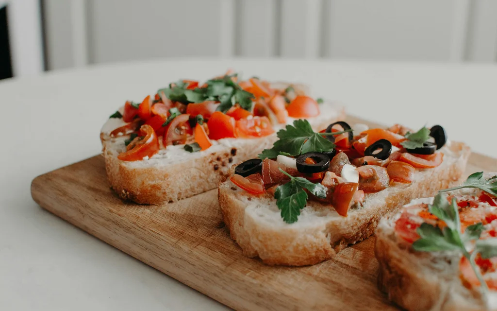 NILS am See Genussmomente: Frühstück nahe Neusiedl am See Drei belegte Brotscheiben mit Tomaten, Oliven und Kräutern auf Holzbrett