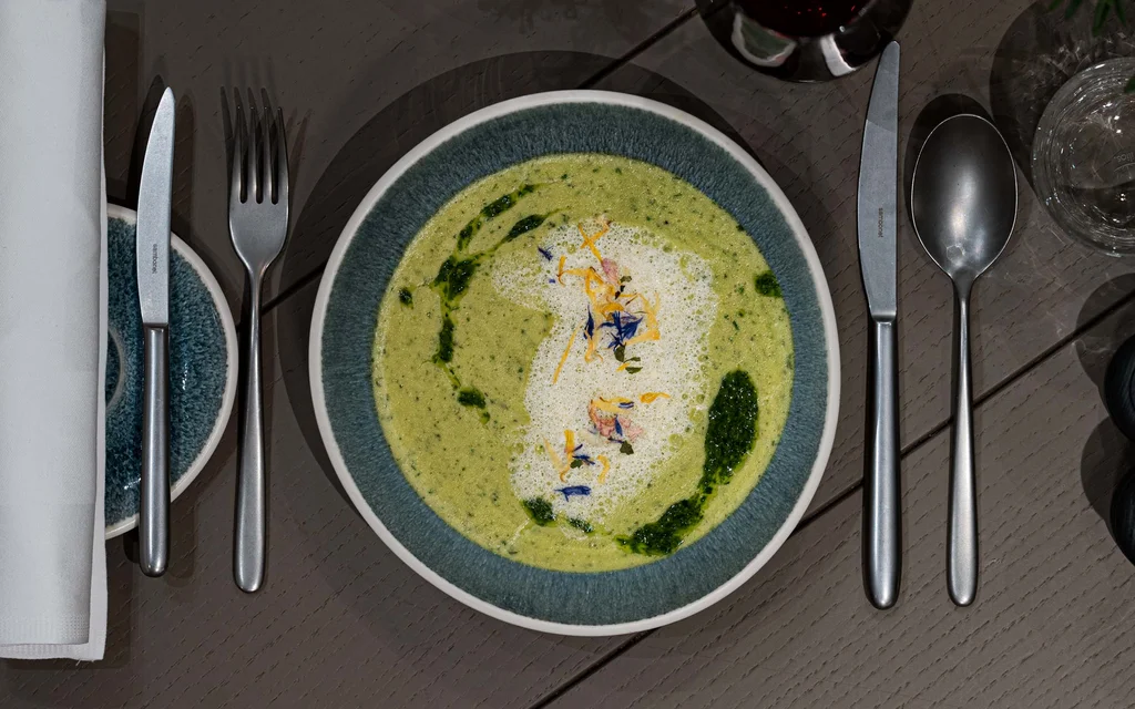 NILS am See | Silvester am Neusiedler See Grüne Suppe mit Schaum und Kräutern auf blauem Teller, Besteck und Glas daneben