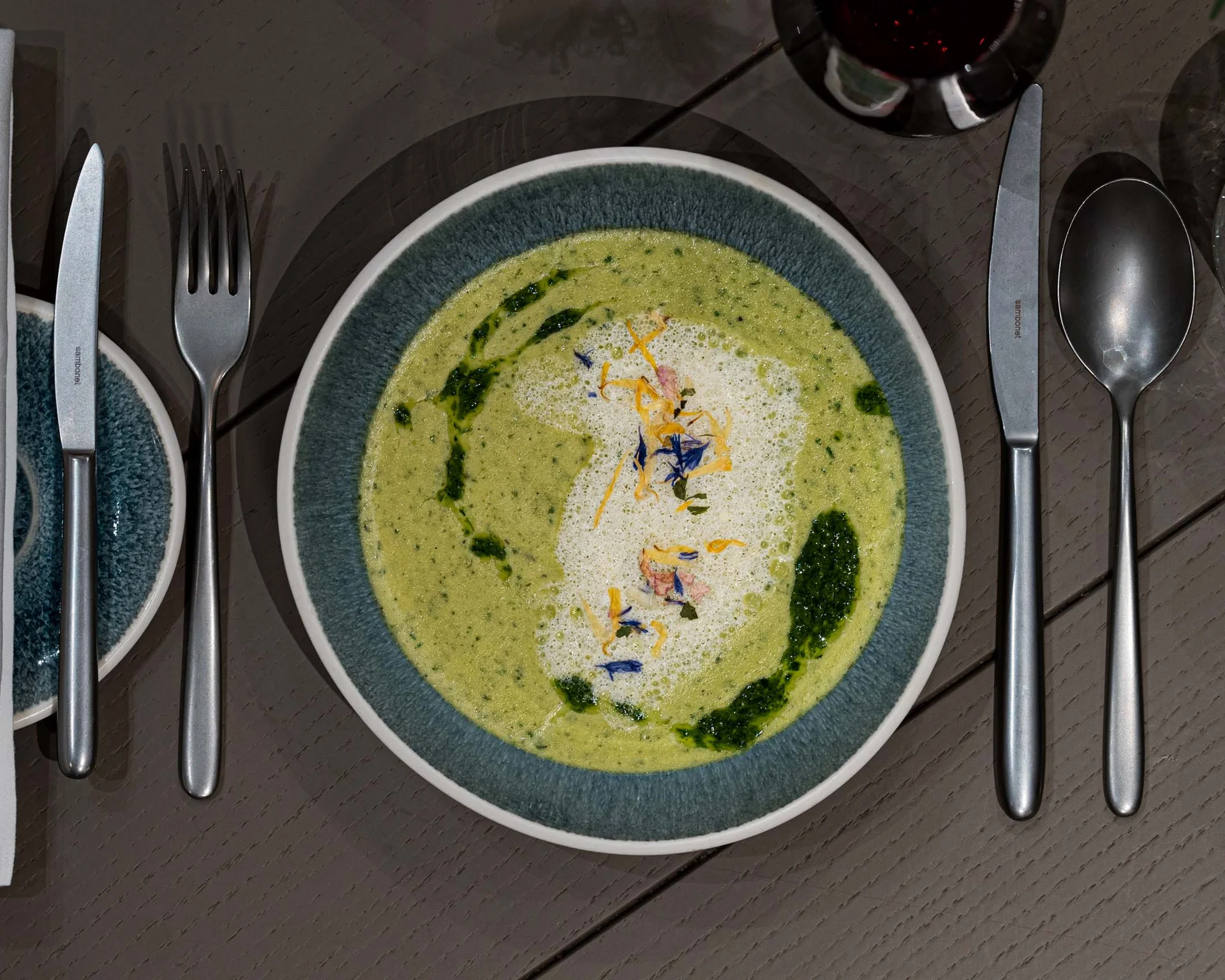 NILS am See: Impressionen eines Orts für das süße Leben Grüne Suppe mit Schaum und Kräutern auf blauem Teller, Besteck und Glas daneben