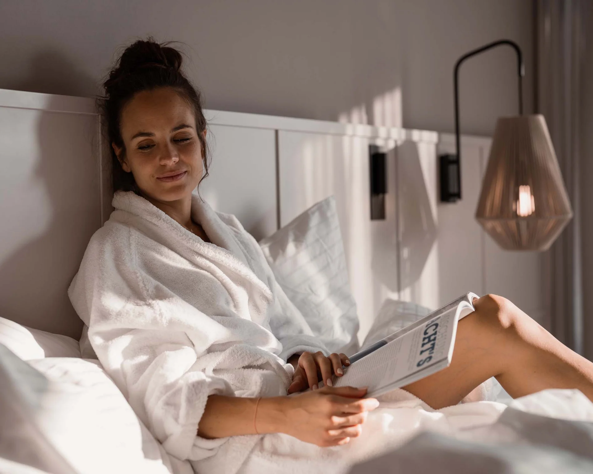 NILS am See: Impressionen eines Orts für das süße Leben Frau in weißem Bademantel liest entspannt auf einem Bett bei natürlichem Licht