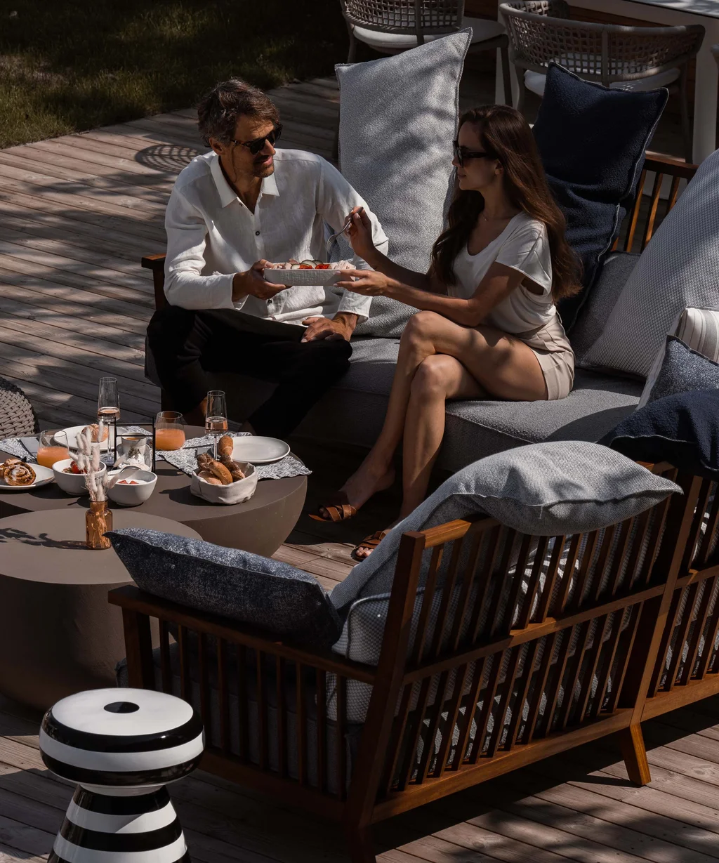 NILS am See Genussmomente: Frühstück nahe Neusiedl am See Paar sitzt auf Outdoor-Sofa und teilt Essen am Holztisch auf der Terrasse.