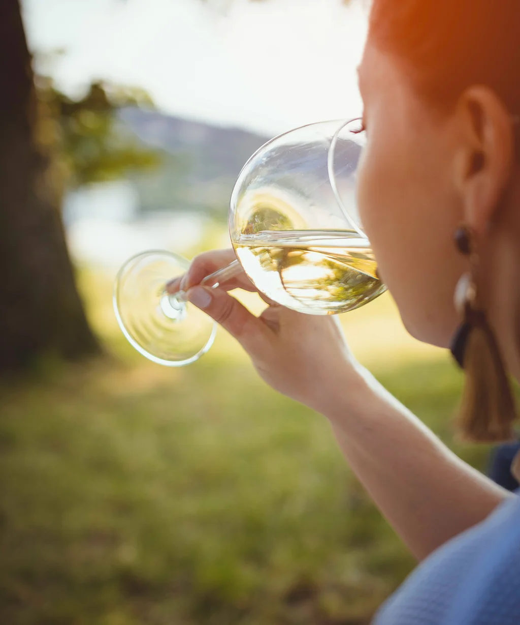 NILS am See: das Boutiquehotel am Neusiedler See Frau trinkt Weißwein draußen bei Sonnenlicht zwischen Bäumen
