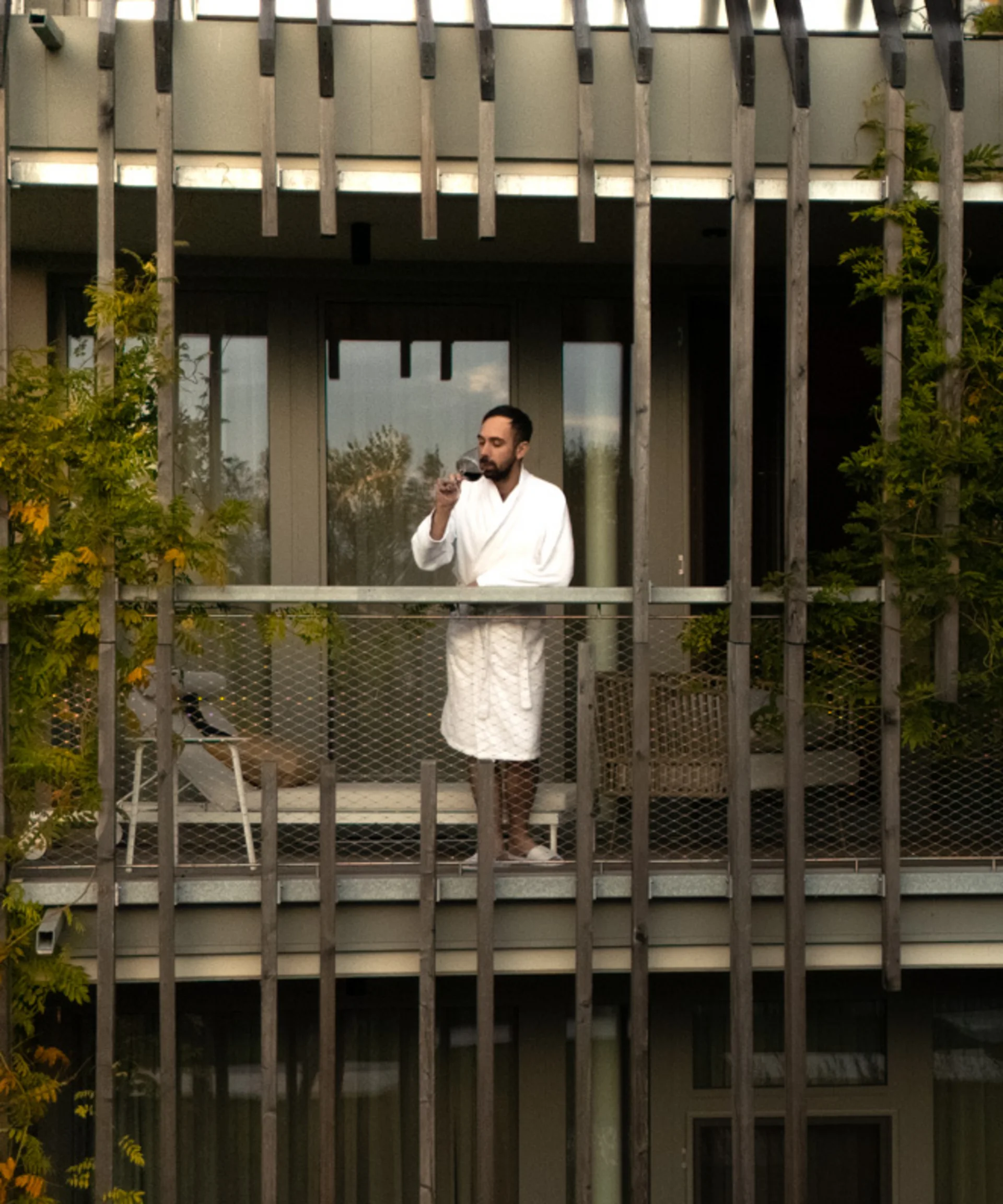 NILS am See: das Boutiquehotel am Neusiedler See Mann im Bademantel trinkt Getränk auf Balkon eines begrünten Gebäudes
