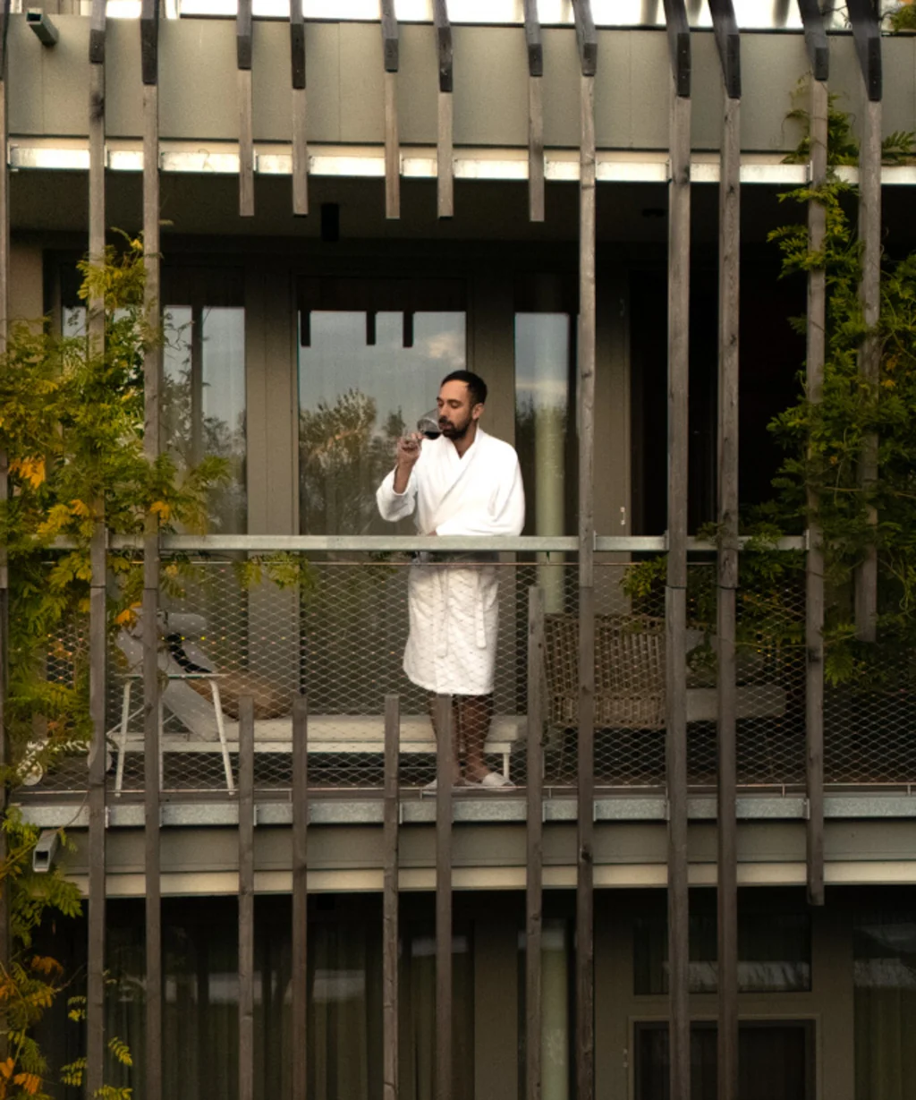 NILS am See: das Boutiquehotel am Neusiedler See Mann im Bademantel trinkt Getränk auf Balkon eines begrünten Gebäudes