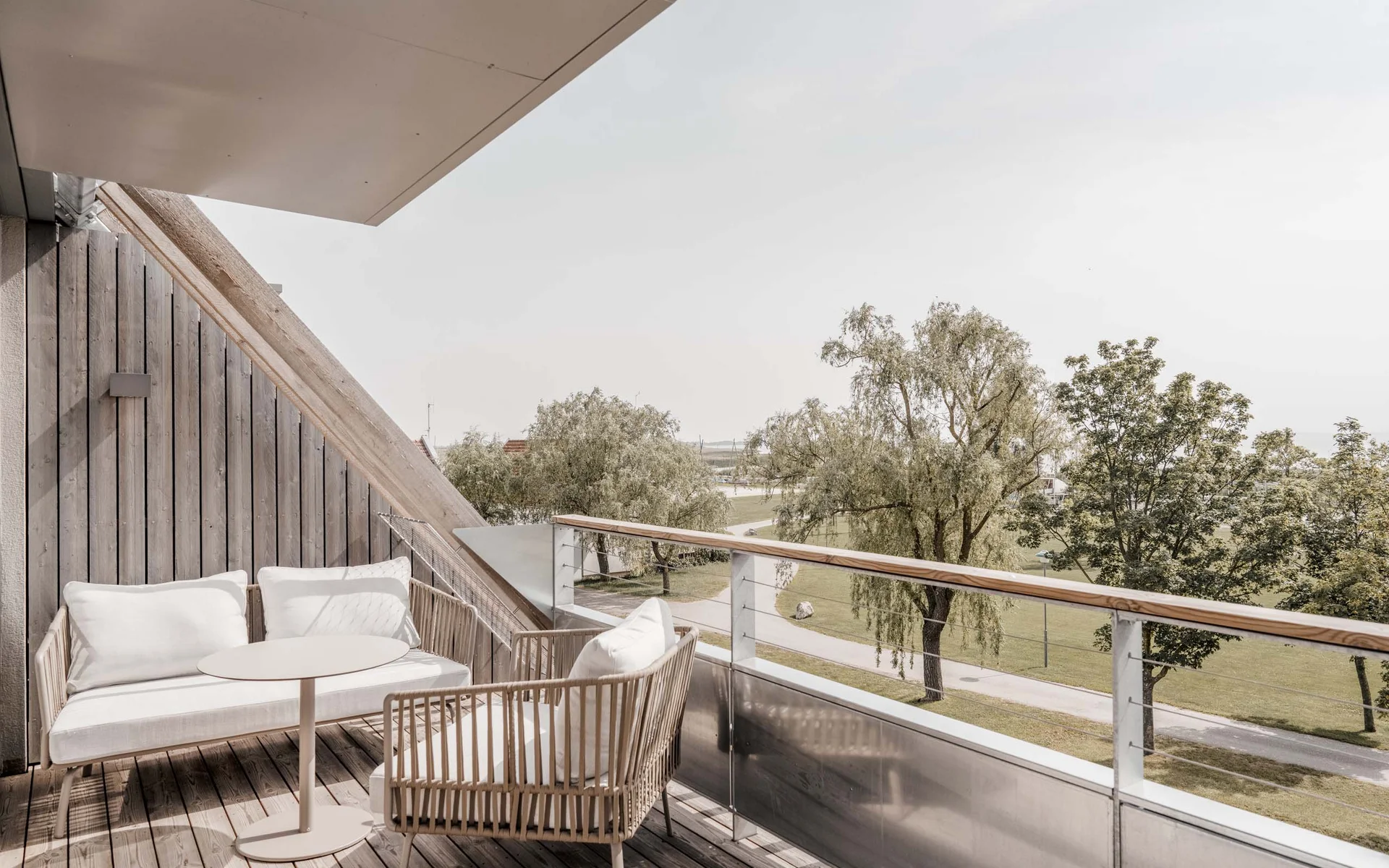 NILS am See: das Boutiquehotel am Neusiedler See Moderner Balkon mit weißen Möbeln und Blick auf grüne Bäume und Weg