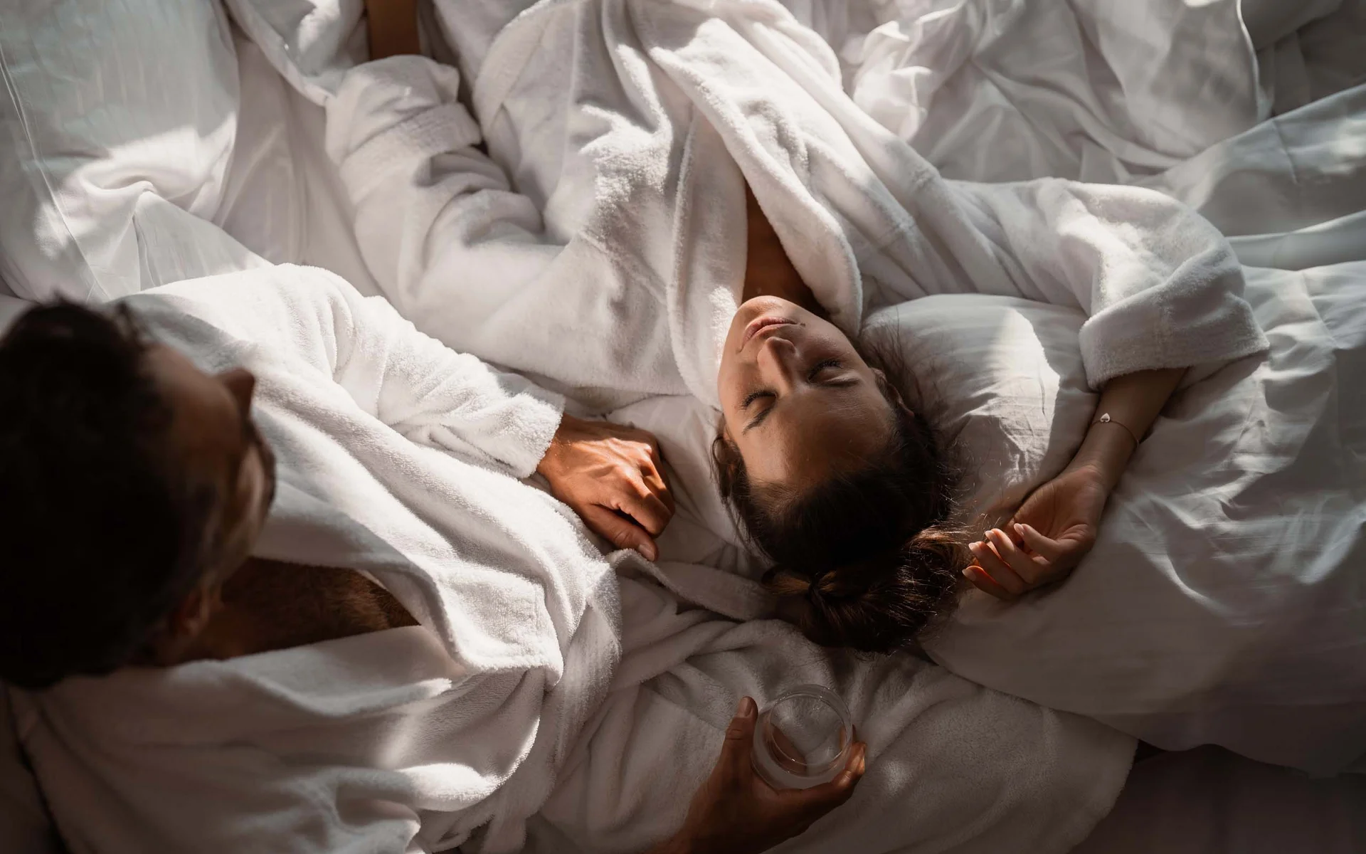 NILS am See: das Boutiquehotel am Neusiedler See Paar in weißen Bademänteln gemütlich im Bett liegend, Frau entspannt