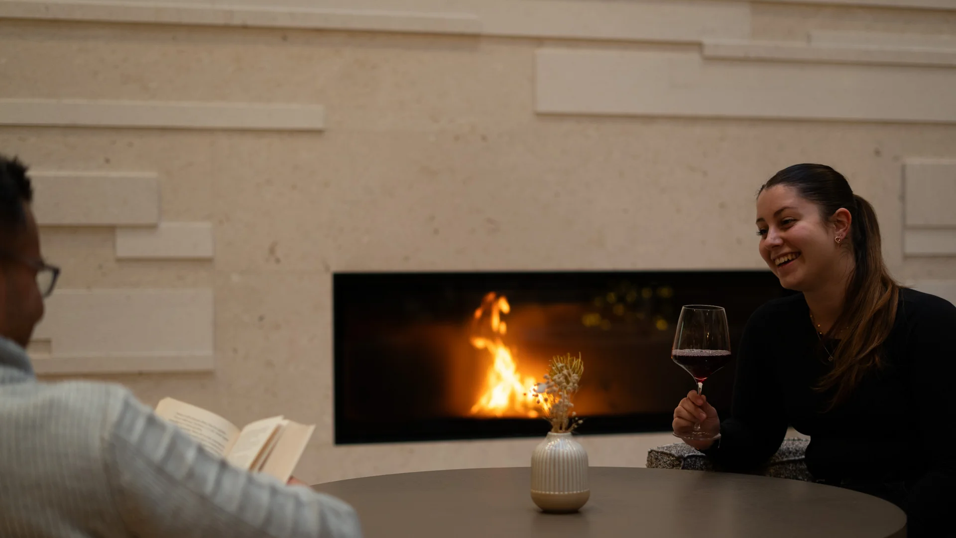 NILS am See: das Boutiquehotel am Neusiedler See Zwei Personen sitzen am Tisch vor einem Kamin, eine liest, die andere trinkt Wein